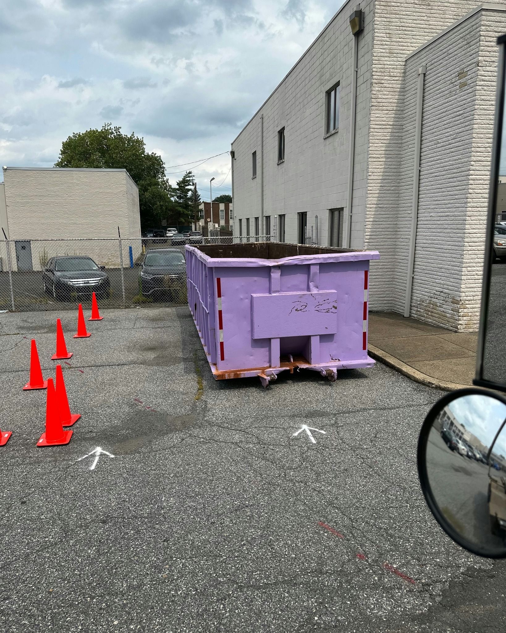 A purple dumpster in a gravel parking lot next to a white building with traffic cones.