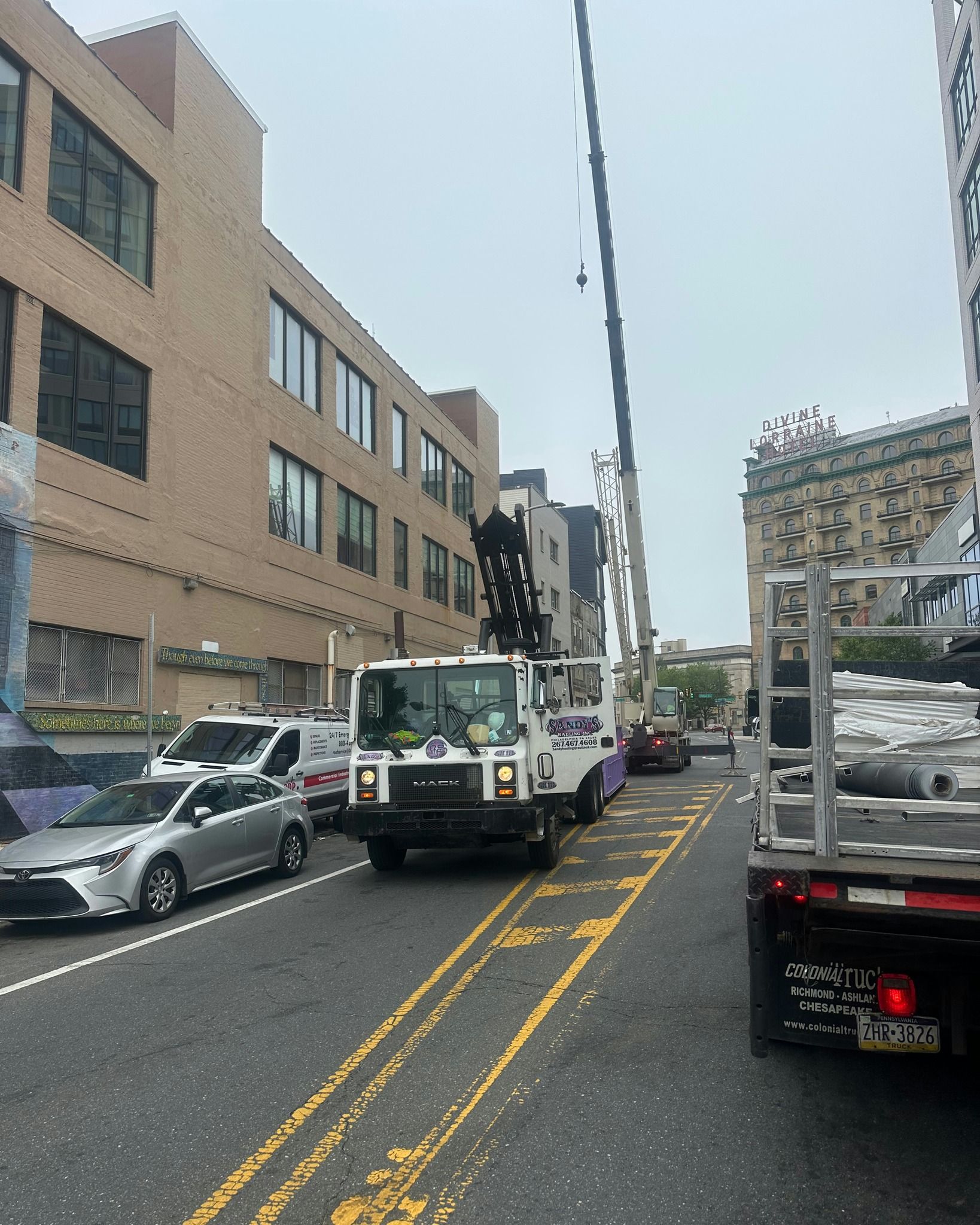 A white garbage truck on a city street with cars. Buildings line the street. Cloudy sky.