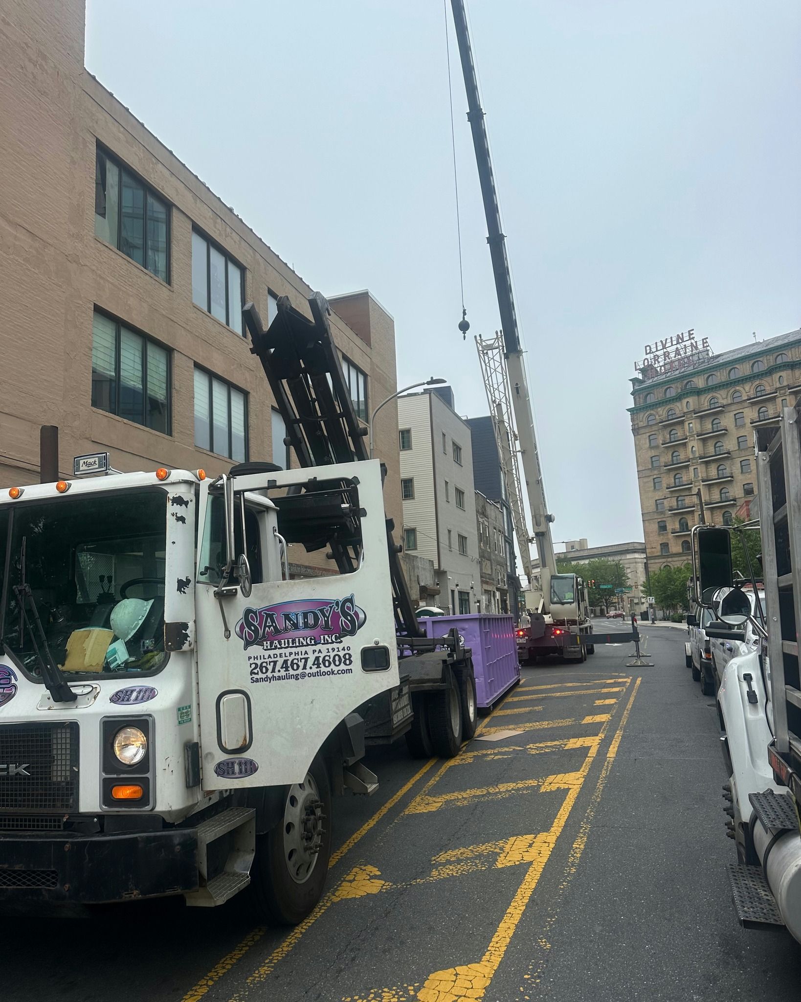 A white truck with a crane on a city street, next to a building under construction.