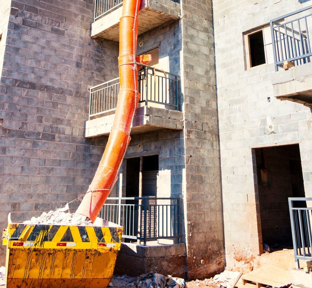 A construction site with a building, a yellow skip, and a chute depositing debris.