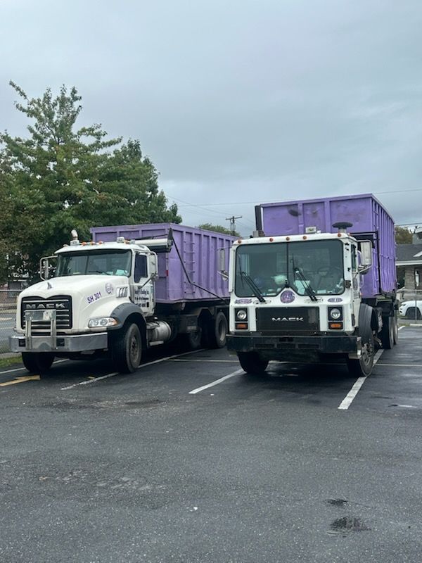 Dump truck with a purple dumpster, parked on a residential street, in front of a multi-story house.