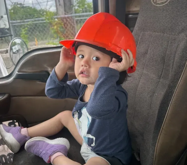 Child in orange hard hat sitting in truck seat, looking surprised.