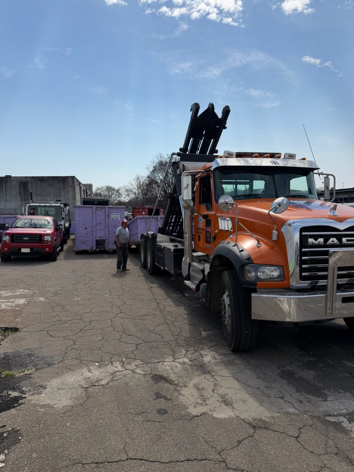 A large purple dumpster next to a building receiving debris via a black chute, on a rainy street.