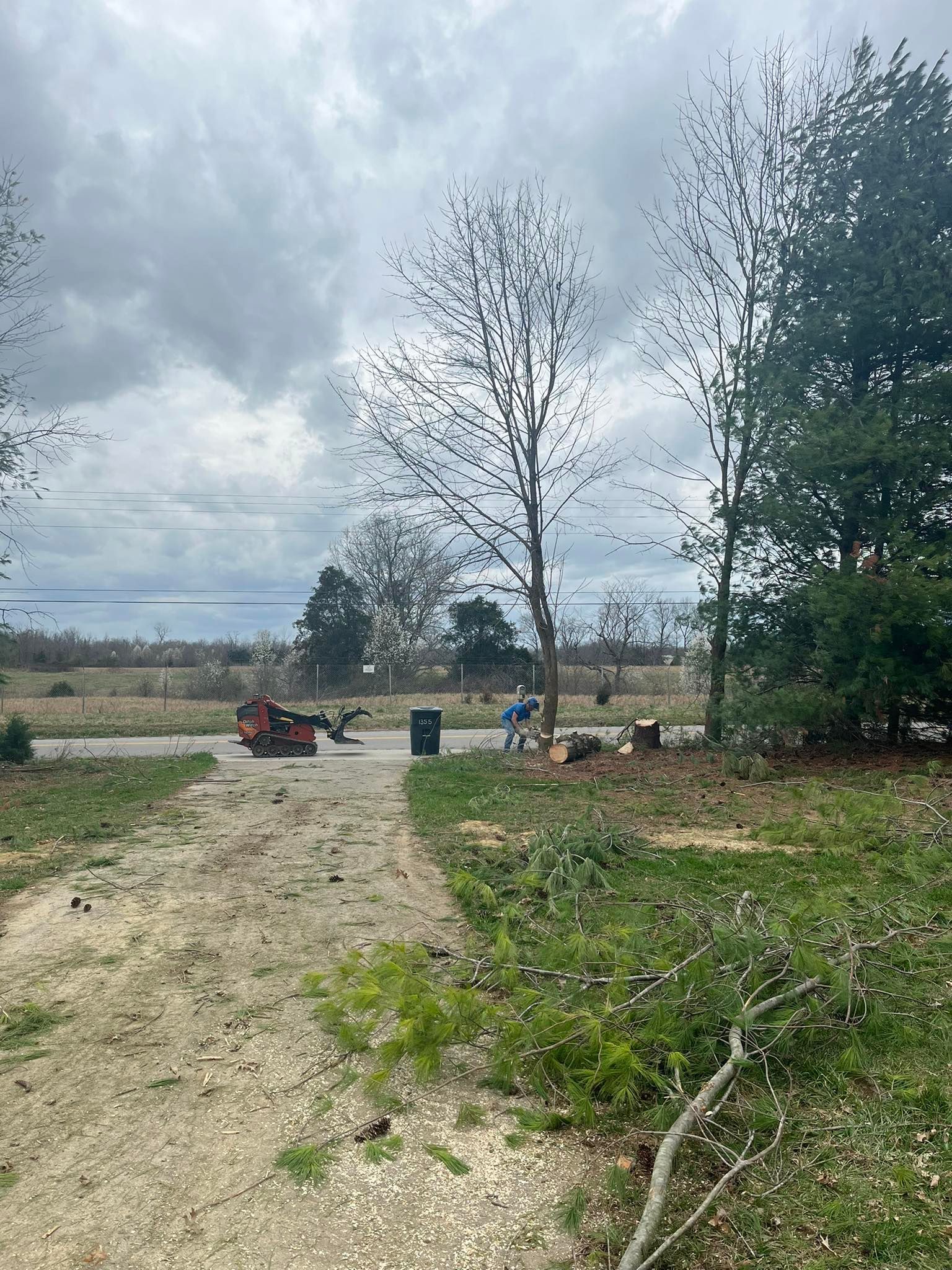 A dirt path leads to a rural field with trees, equipment, and people under a cloudy sky. A dirt path leads to a rural field with trees, equipment, and people under a cloudy sky.