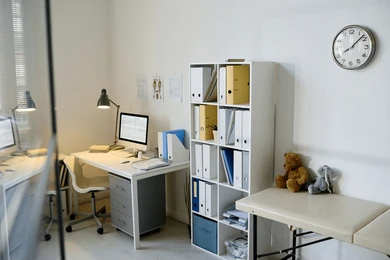 A doctor 's office with two desks , a shelf , a clock and a teddy bear.