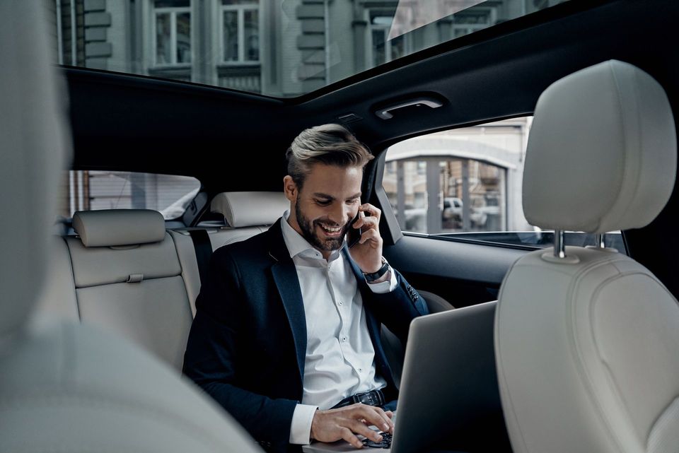 Un uomo è seduto sul sedile posteriore di un'auto e sta usando un computer portatile e parlando al cellulare.