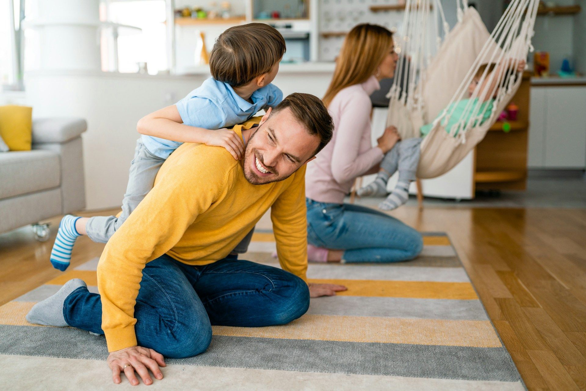 A man is kneeling on the floor with two children and a hammock in the background.