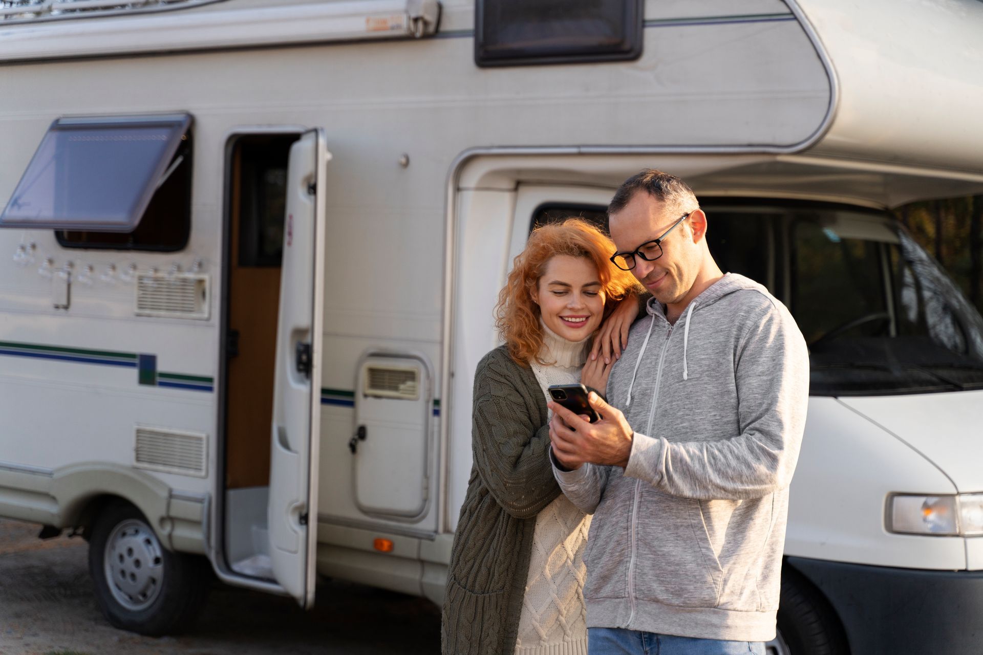 A man and a woman are standing in front of a camper van looking at a cell phone.