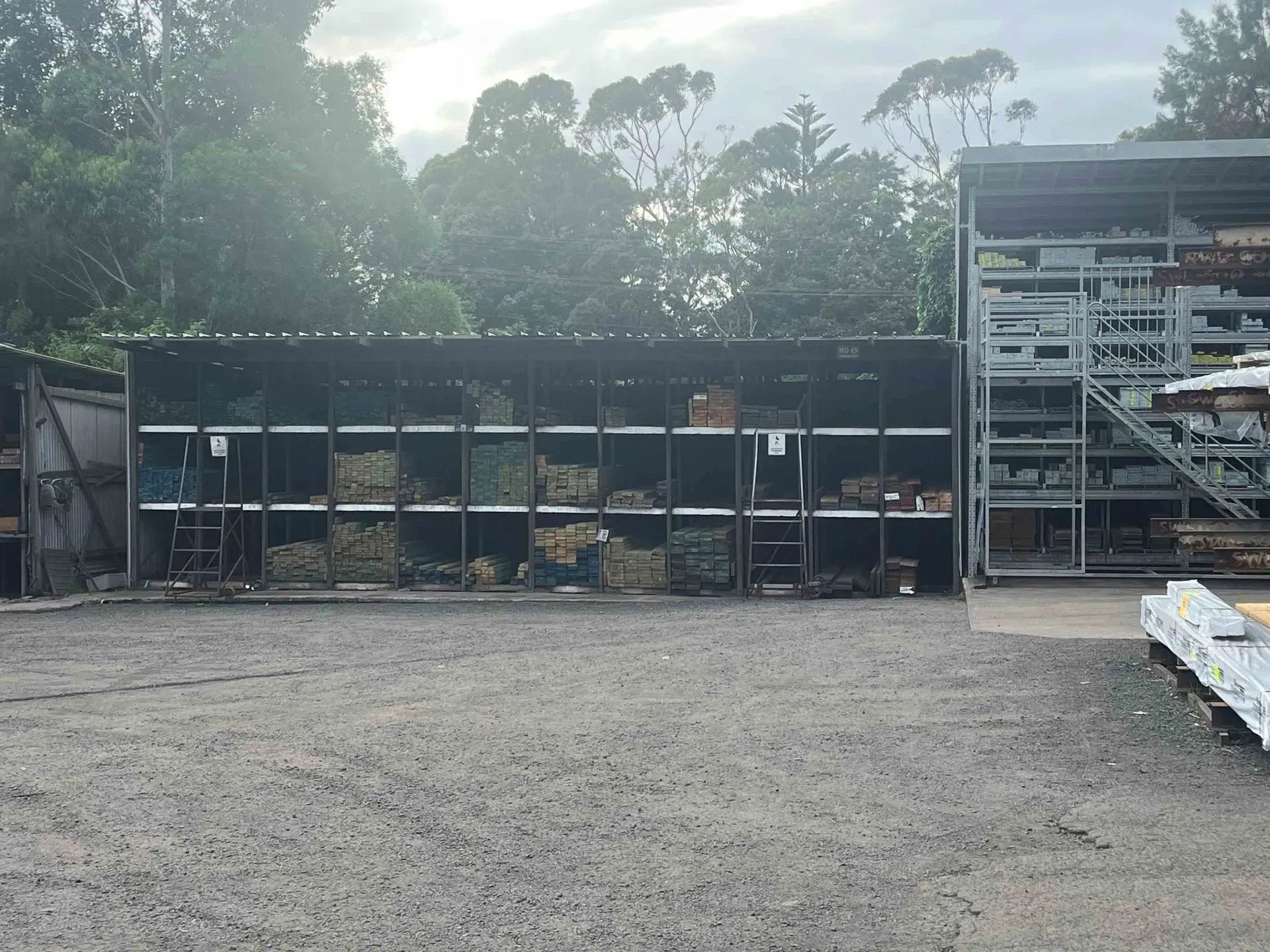 Stacks of light-colored sawn wooden planks in the foreground with a pile of unprocessed logs in the background.