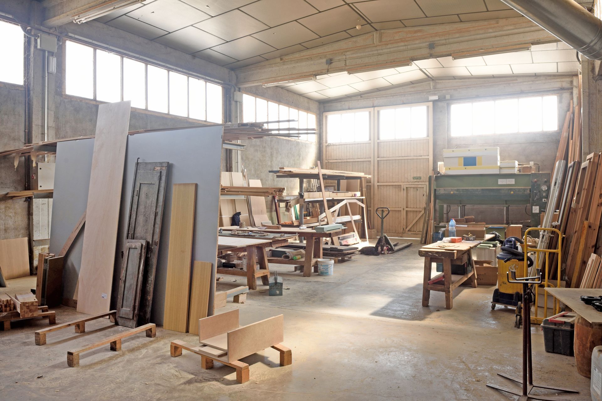 A bright, industrial woodworking workshop filled with lumber, workbenches, and tools under a high, paneled ceiling. A bright, industrial woodworking workshop filled with lumber, workbenches, and tools under a high, paneled ceiling.