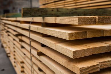 A large stack of light-colored, rough-cut lumber boards stored in a warehouse.