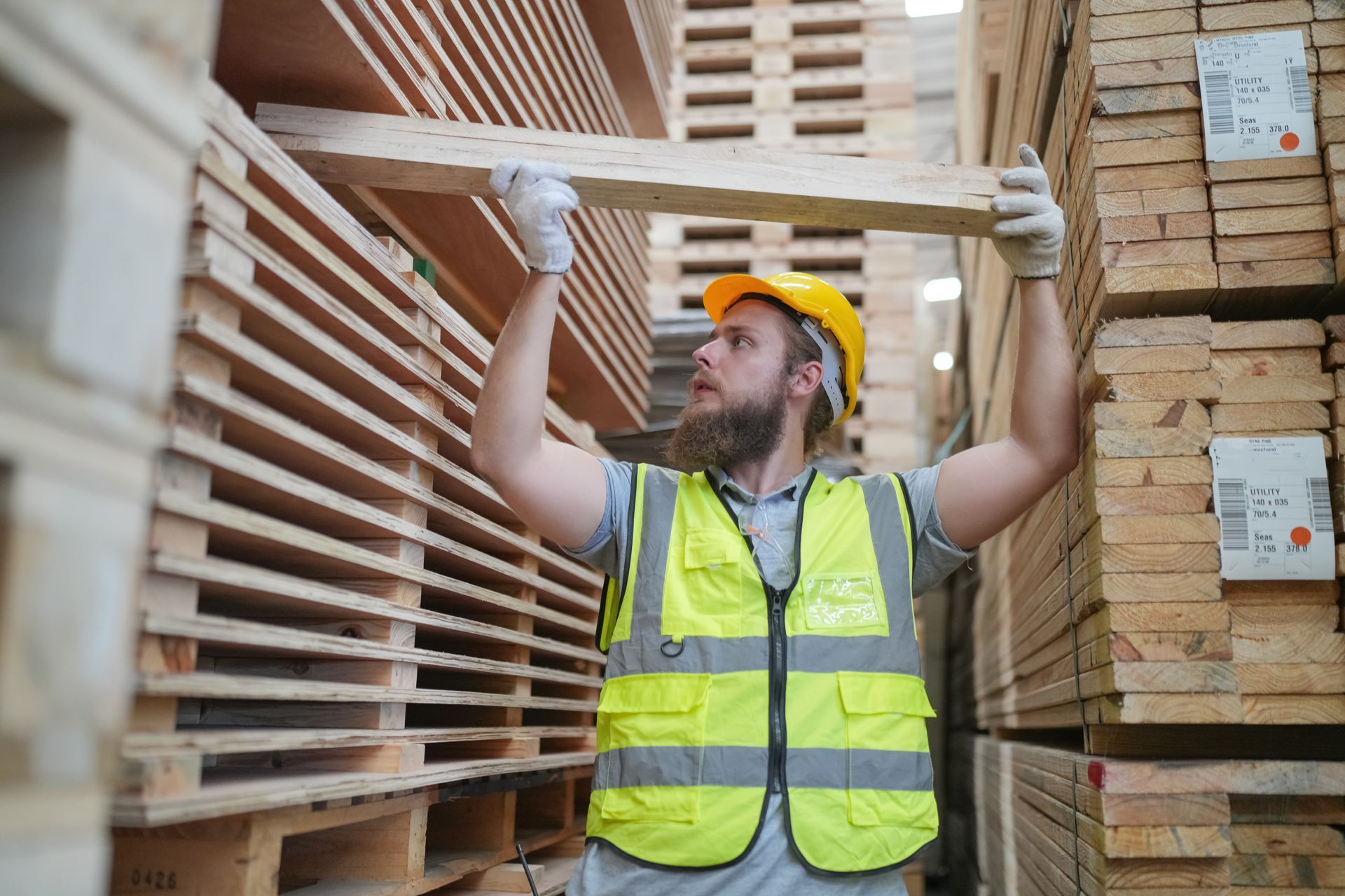 A warehouse worker in a yellow hard hat and high-visibility vest lifts a wooden board while working near stacked lumber. A warehouse worker in a yellow hard hat and high-visibility vest lifts a wooden board while working near stacked lumber.