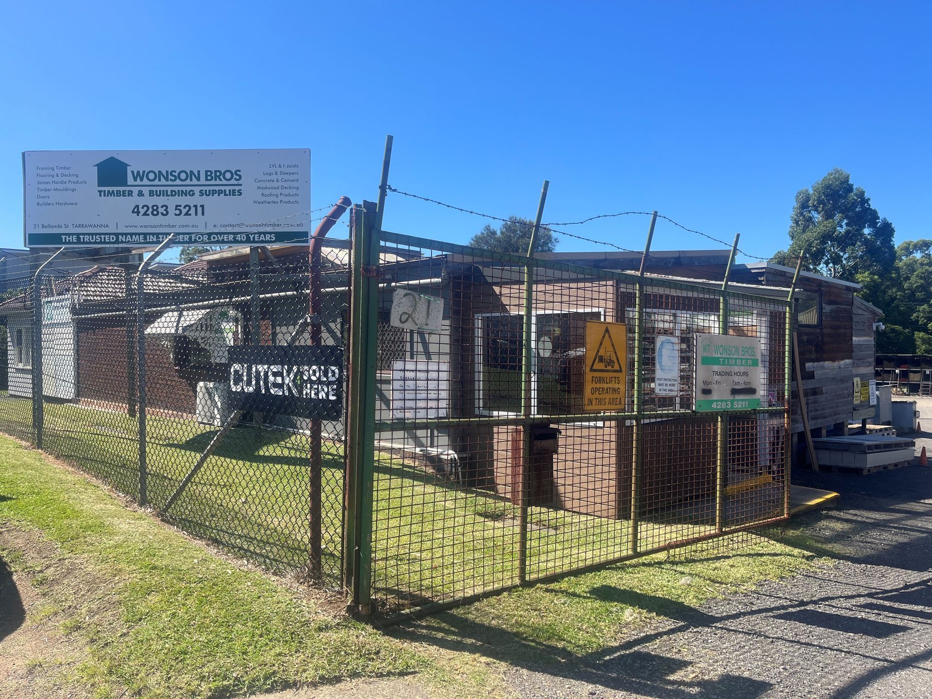 A fenced brick building and storage yard under a clear blue sky, featuring a large white business sign and security wire.