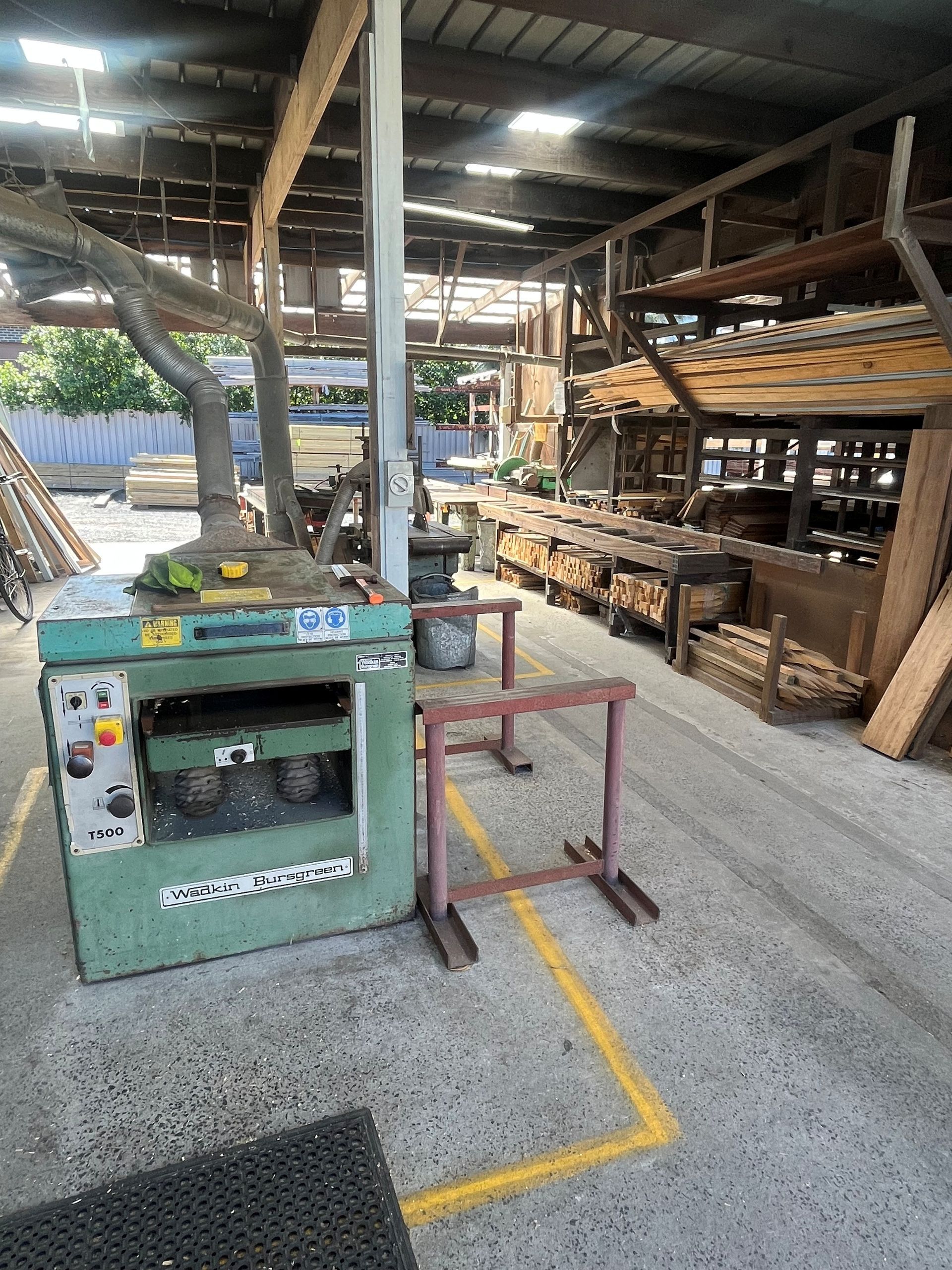 Green industrial planer in a woodworking shop with stacks of lumber and workshop tools in the background.