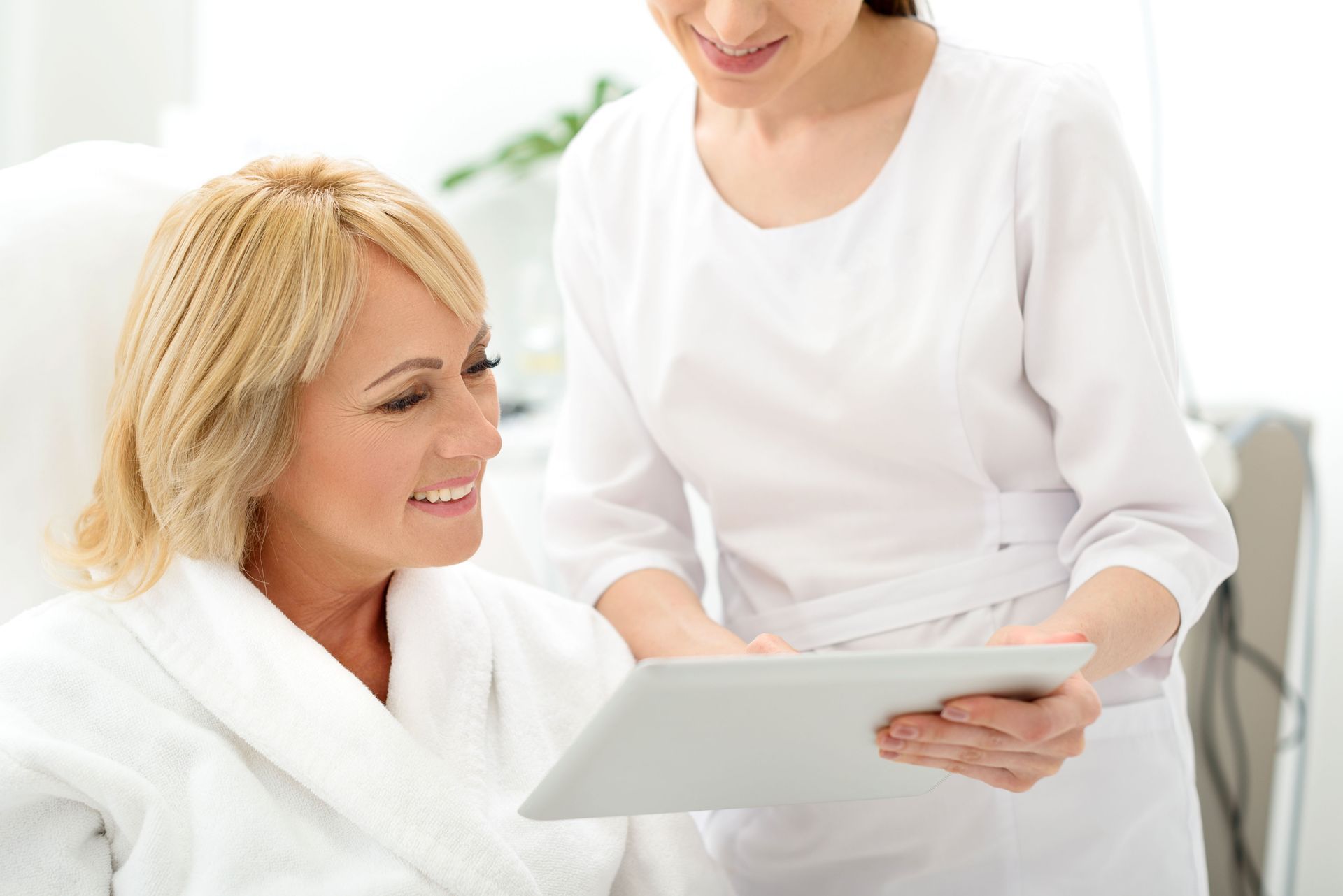 Woman in bathrobe consulting with a person holding a tablet, smiling. White setting.