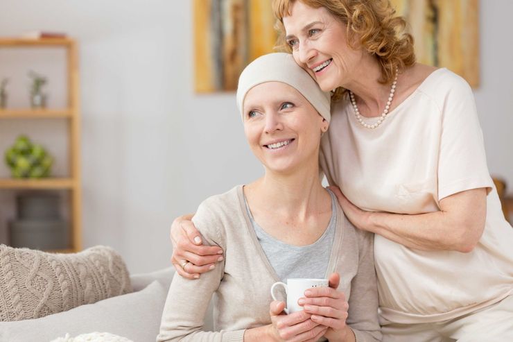 Woman with headscarf, smiling, holding a cup, embraced by another woman; warm colors, interior setting.