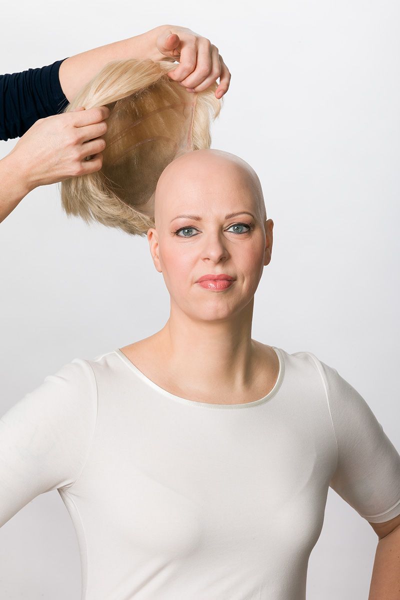 Person with a shaved head having a wig placed on top by someone's hands. White background.