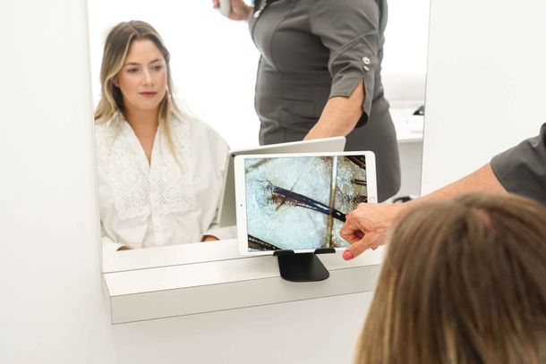Woman in mirror, hair examined with tablet. Person points to image of scalp. White setting.