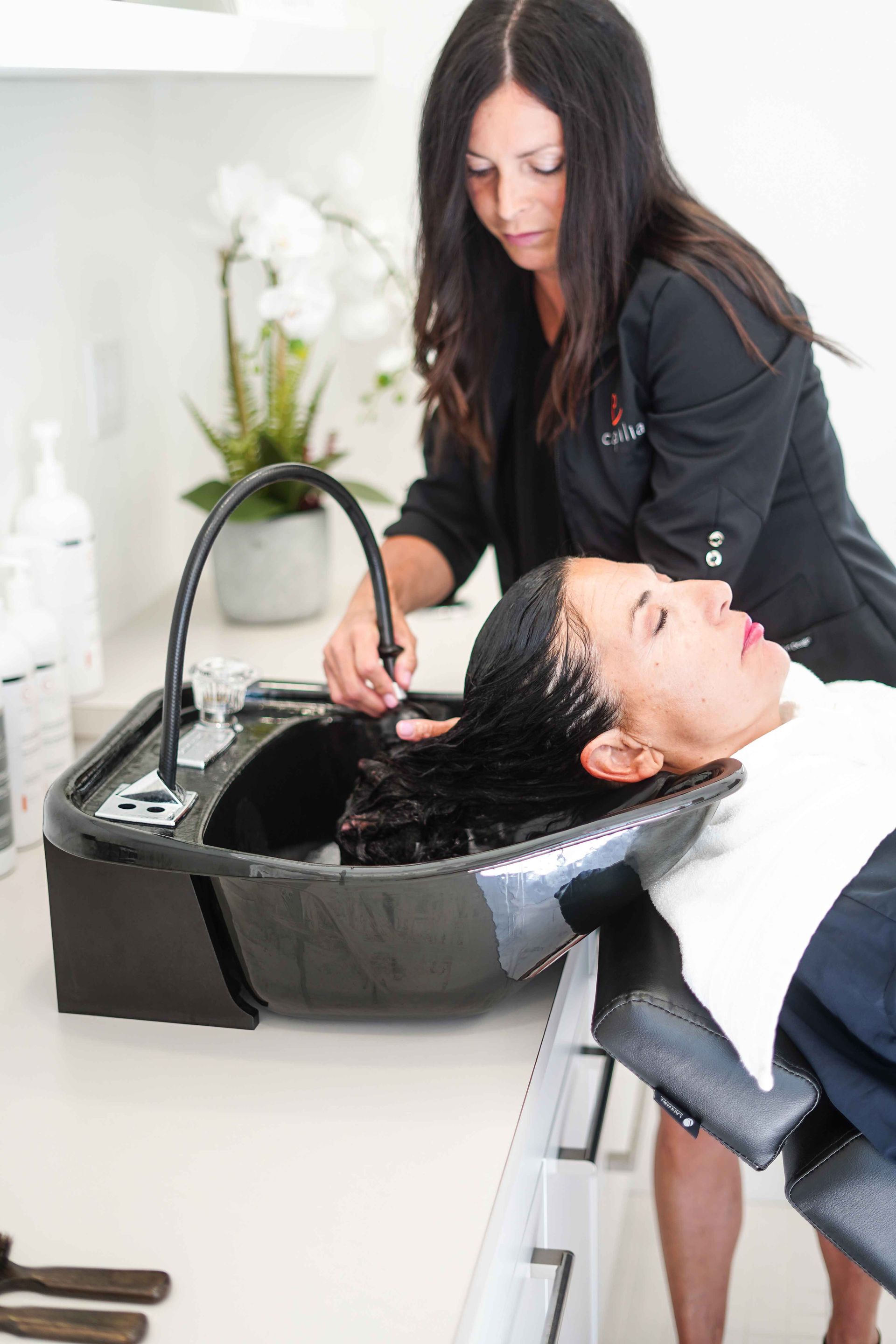 Hairdresser washing a client's hair in a salon basin. The client reclines, the hairdresser holds the nozzle.