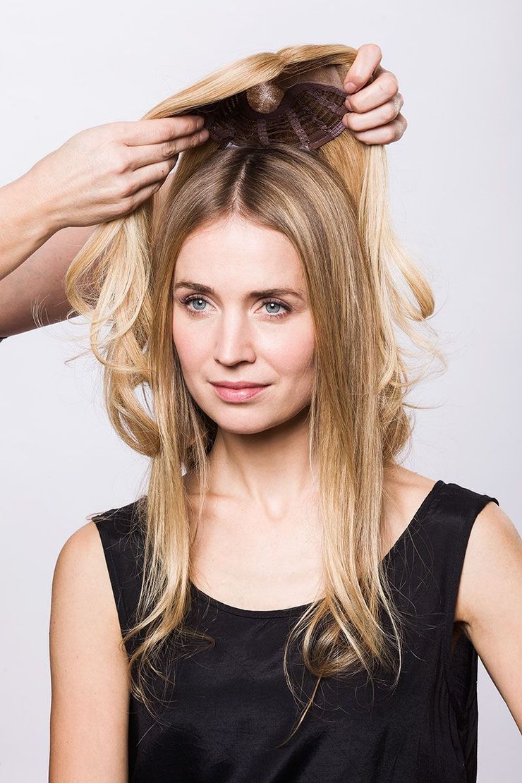 Woman receiving a hairpiece installation, hands adjusting it. Blonde hair, black top, studio setting.