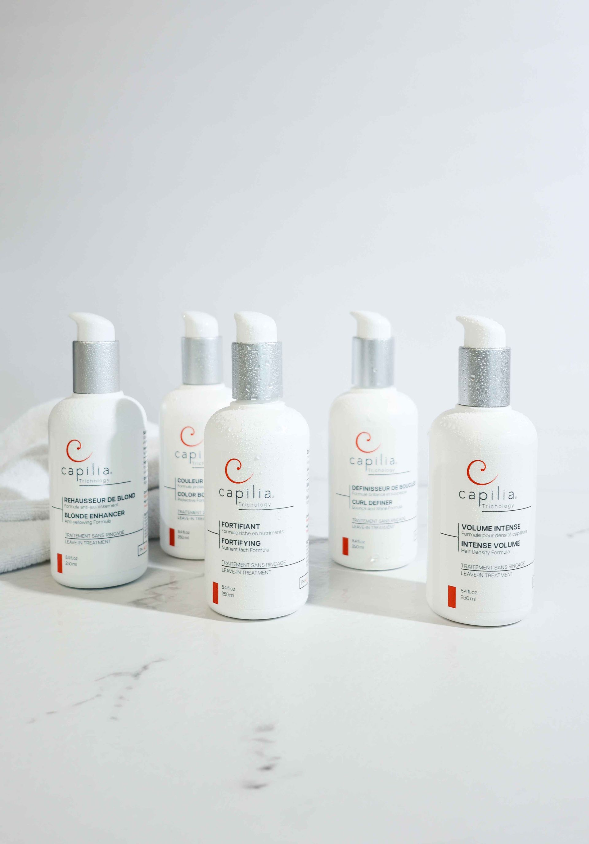 Cinq flacons cosmétiques à pompe blancs identiques, avec des bouchons argentés, disposés en rangée sur une surface en marbre blanc.