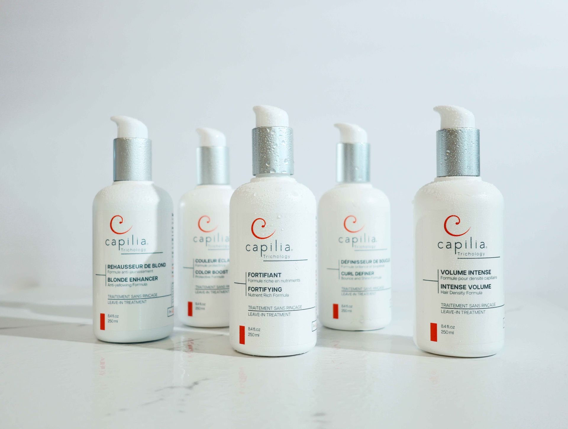 Cinq flacons-pompes blancs de produits capillaires Capilia, alignés sur une surface blanche.
