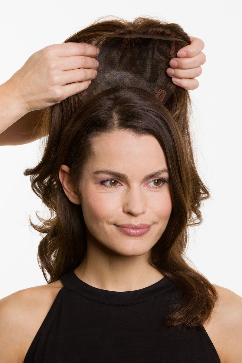 Person trying on a hairpiece. Another person's hands hold the piece above her head. She smiles, and is wearing a black top.