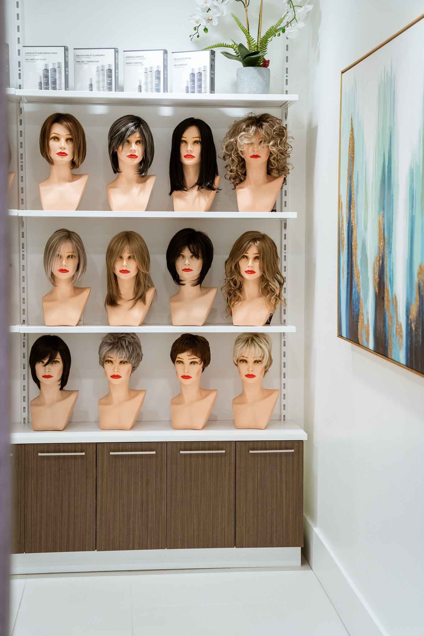Wigs displayed on mannequins on shelves in a beauty salon. Brown cabinet below, artwork on the wall.