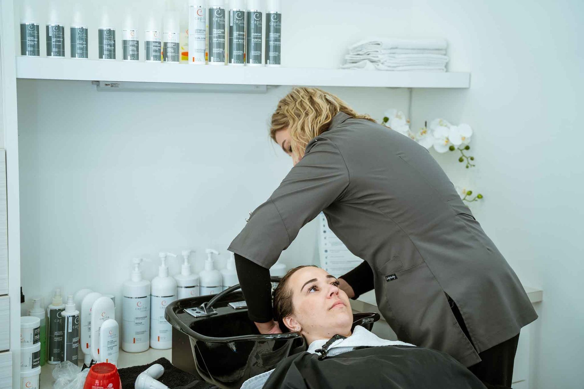 Une coiffeuse lave les cheveux d'une cliente au lavabo d'un salon, avec diverses bouteilles sur des étagères en arrière-plan.