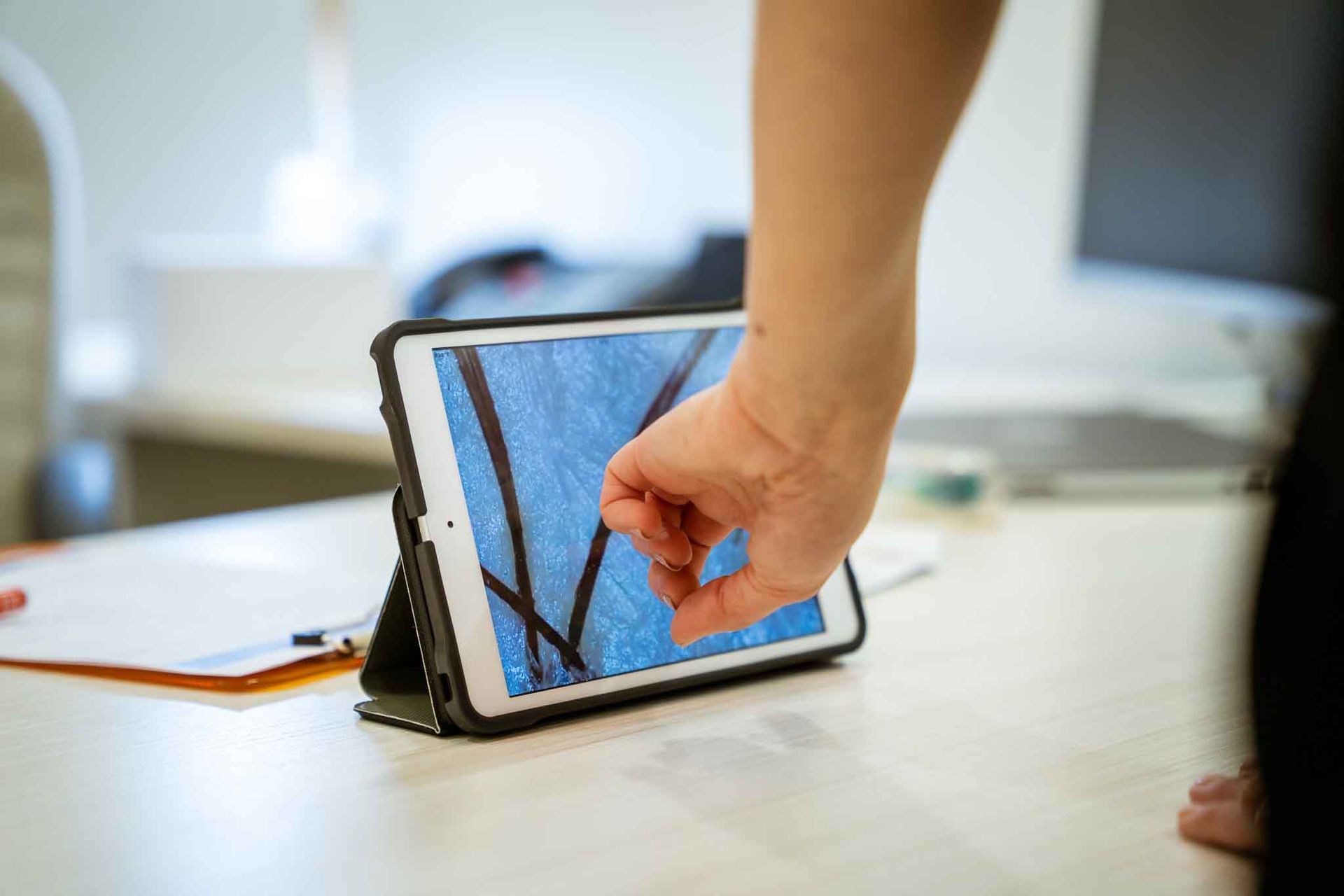 Person pointing at a tablet displaying a close-up of blue lines, on a desk in an office.