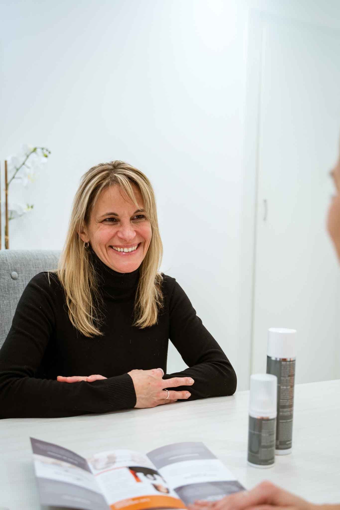 Woman at a table smiles, facing another person.  Two skincare product bottles, a brochure. White room.