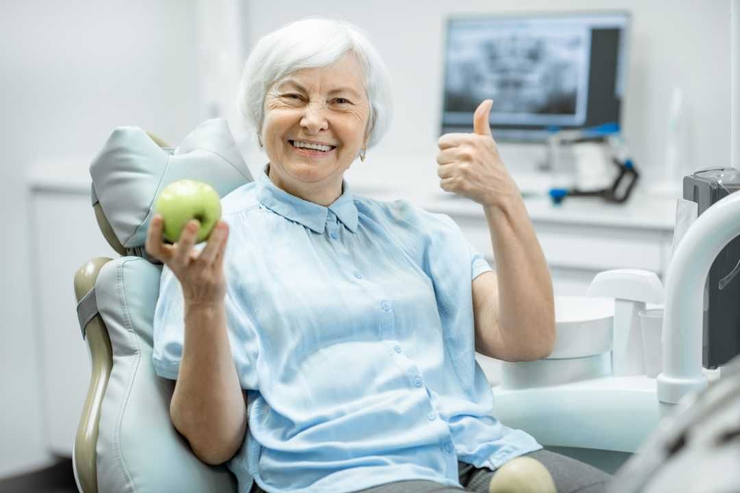 Smiling patient in a dental chair holding an apple and giving a thumbs-up in a bright clinic
