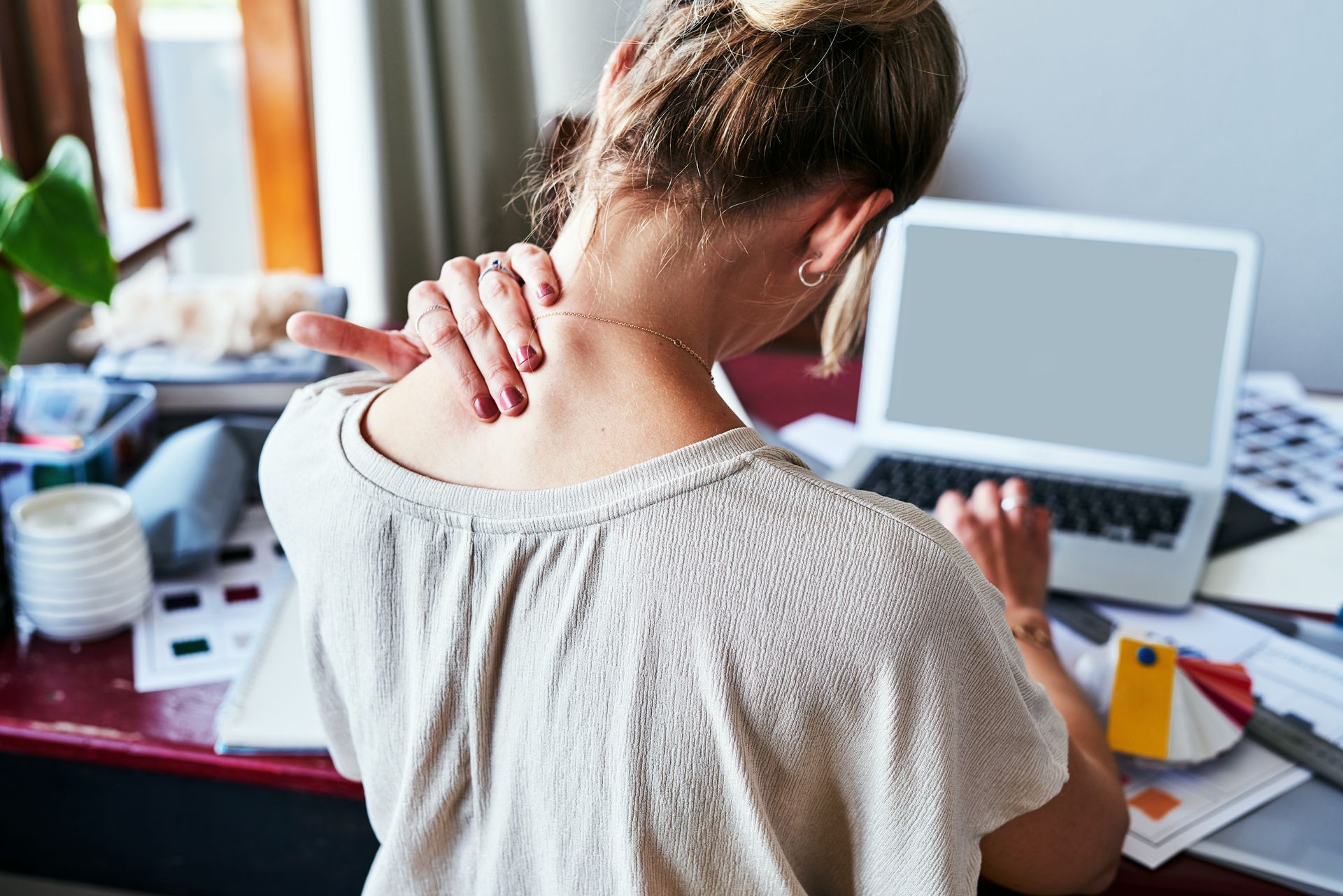 A woman is sitting at a desk with a laptop and holding her neck.