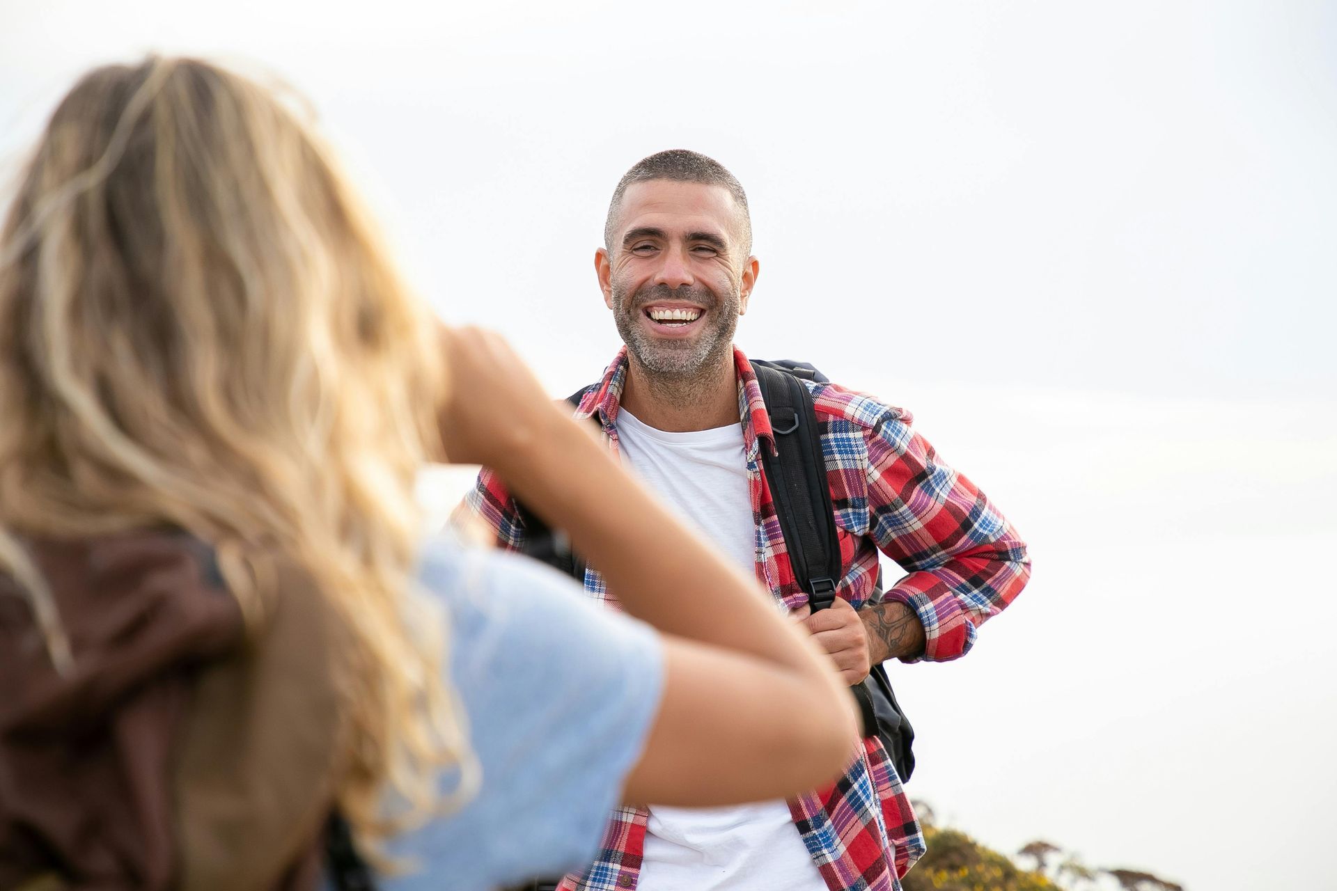 A person with long hair taking a photo of a smiling hiker wearing a red plaid shirt and backpack outdoors.