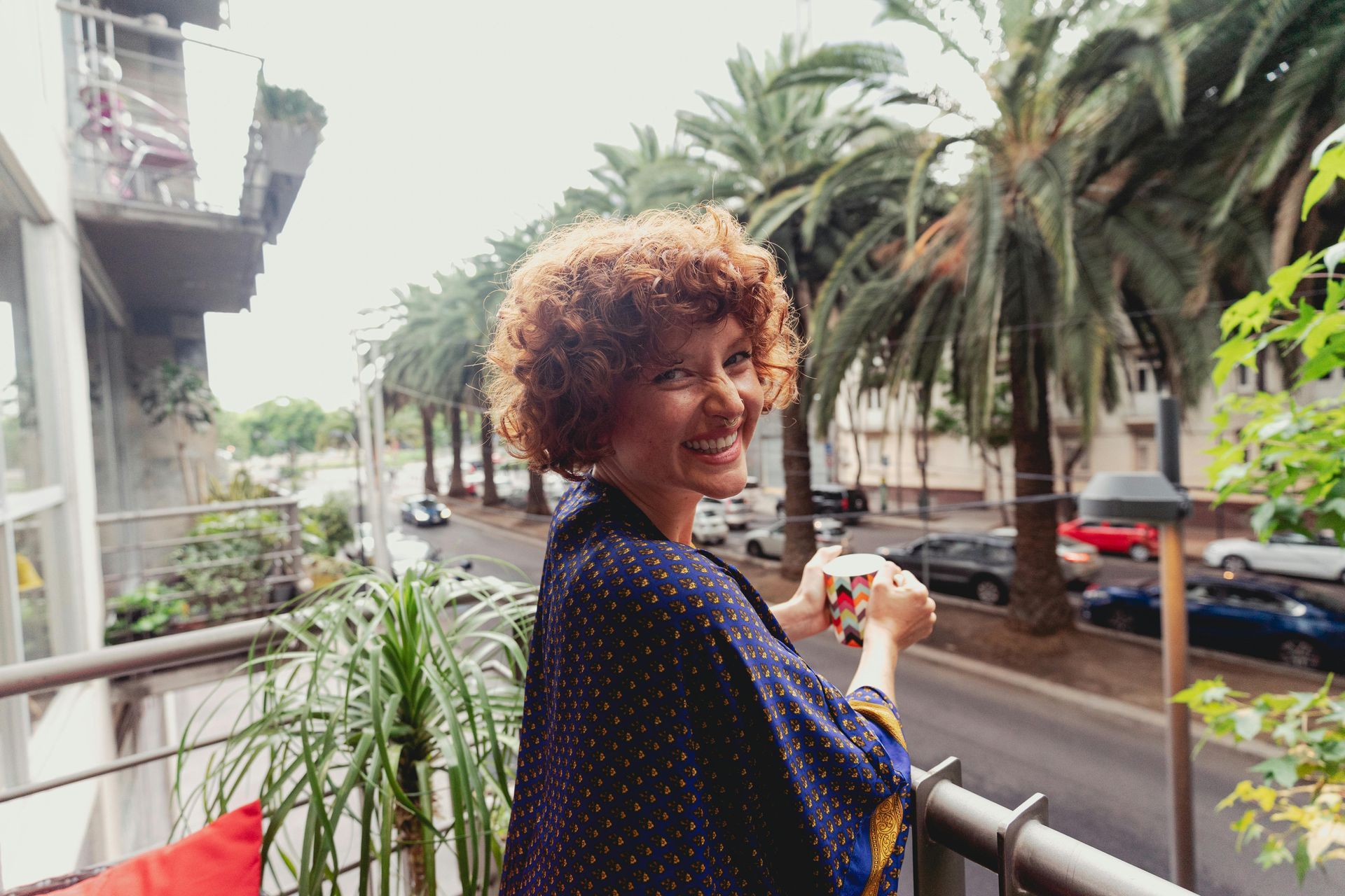 A person with curly hair smiling while holding a mug on a balcony overlooking a palm-lined street.