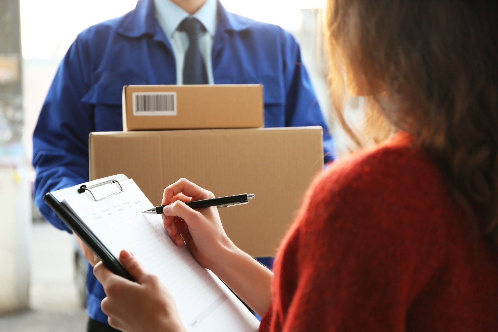 A woman is writing on a clipboard while a delivery man is holding boxes.