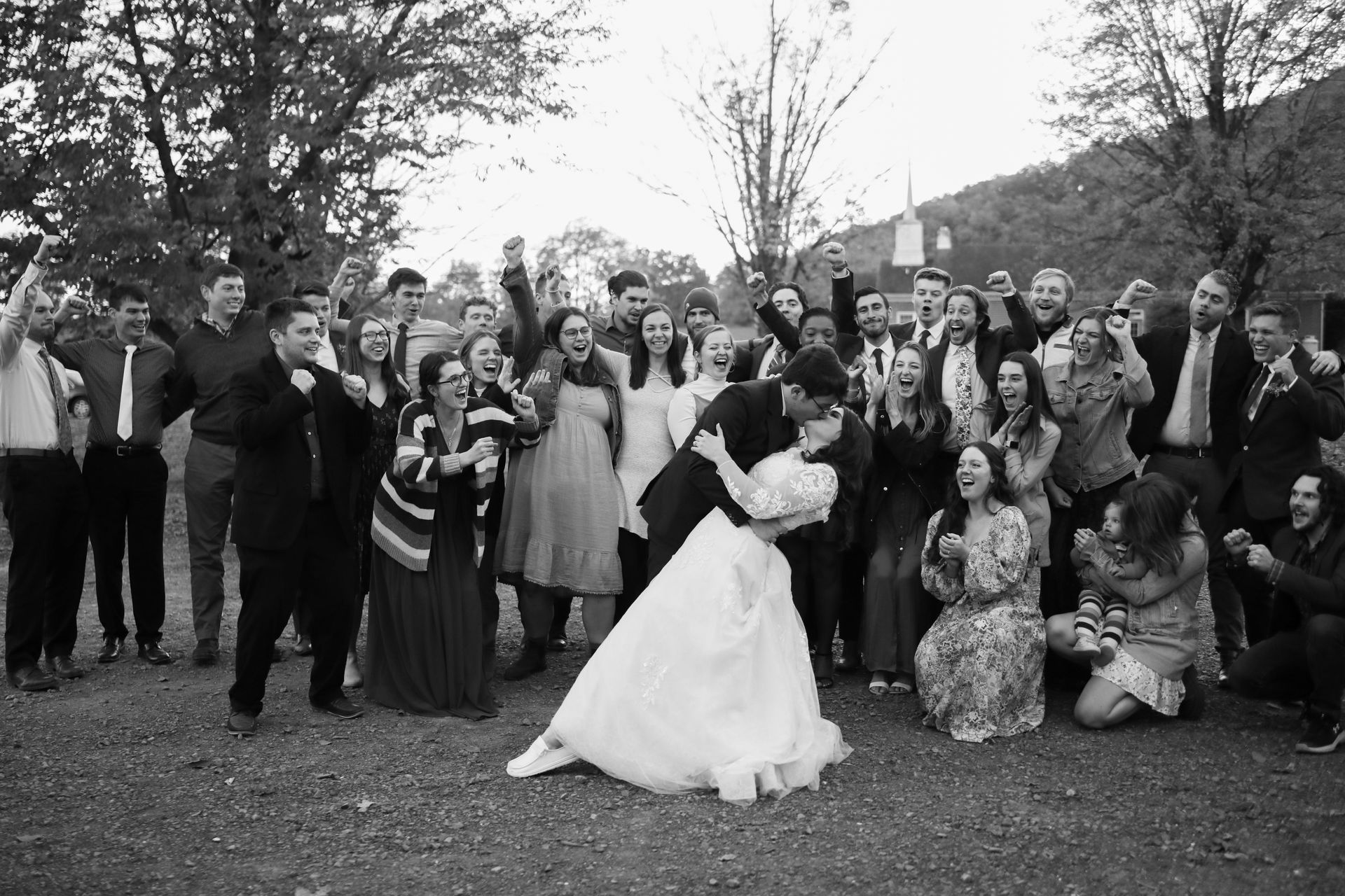 A black and white photo of a bride and groom kissing in front of their wedding party.