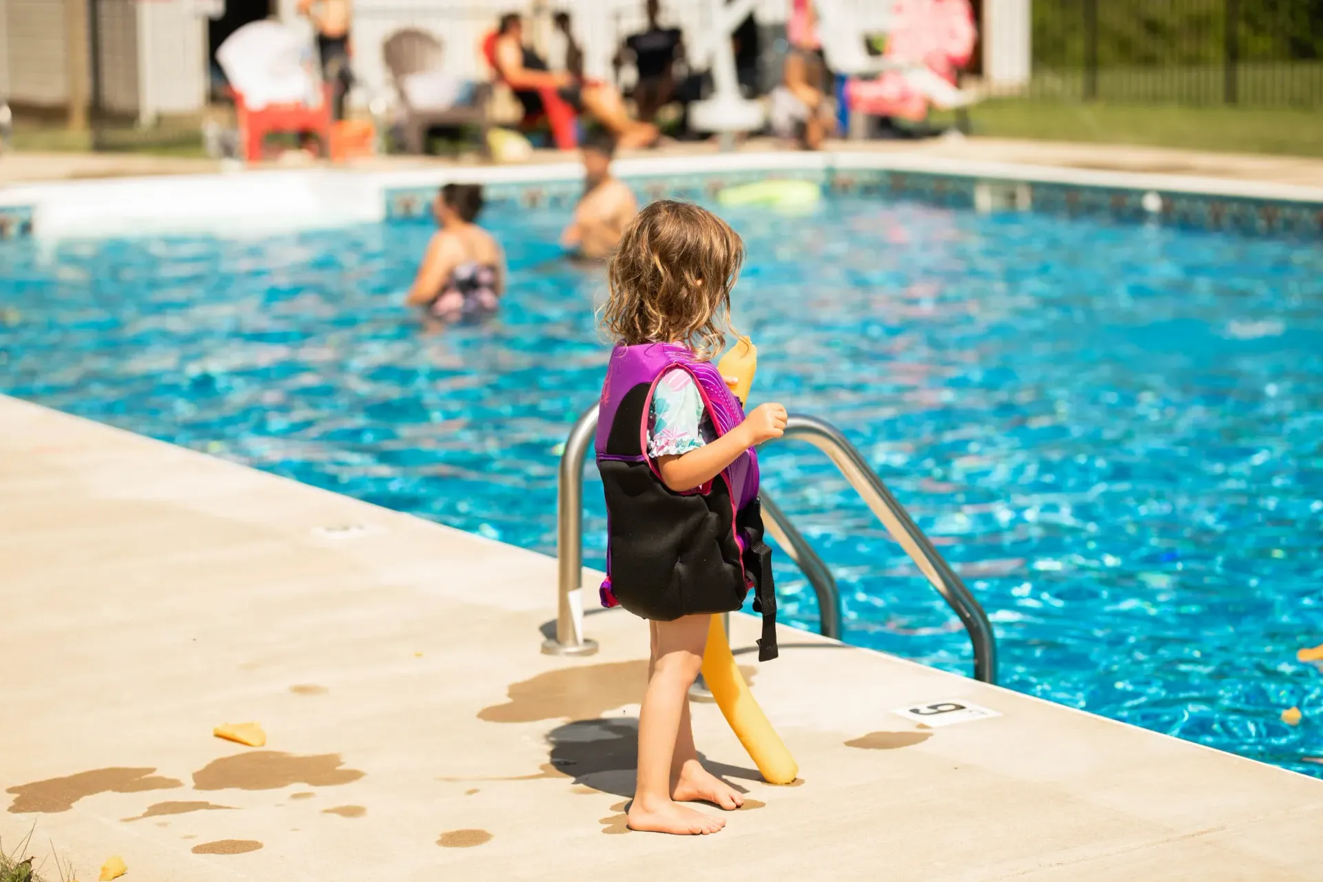 A little girl in a life jacket is standing next to a swimming pool.