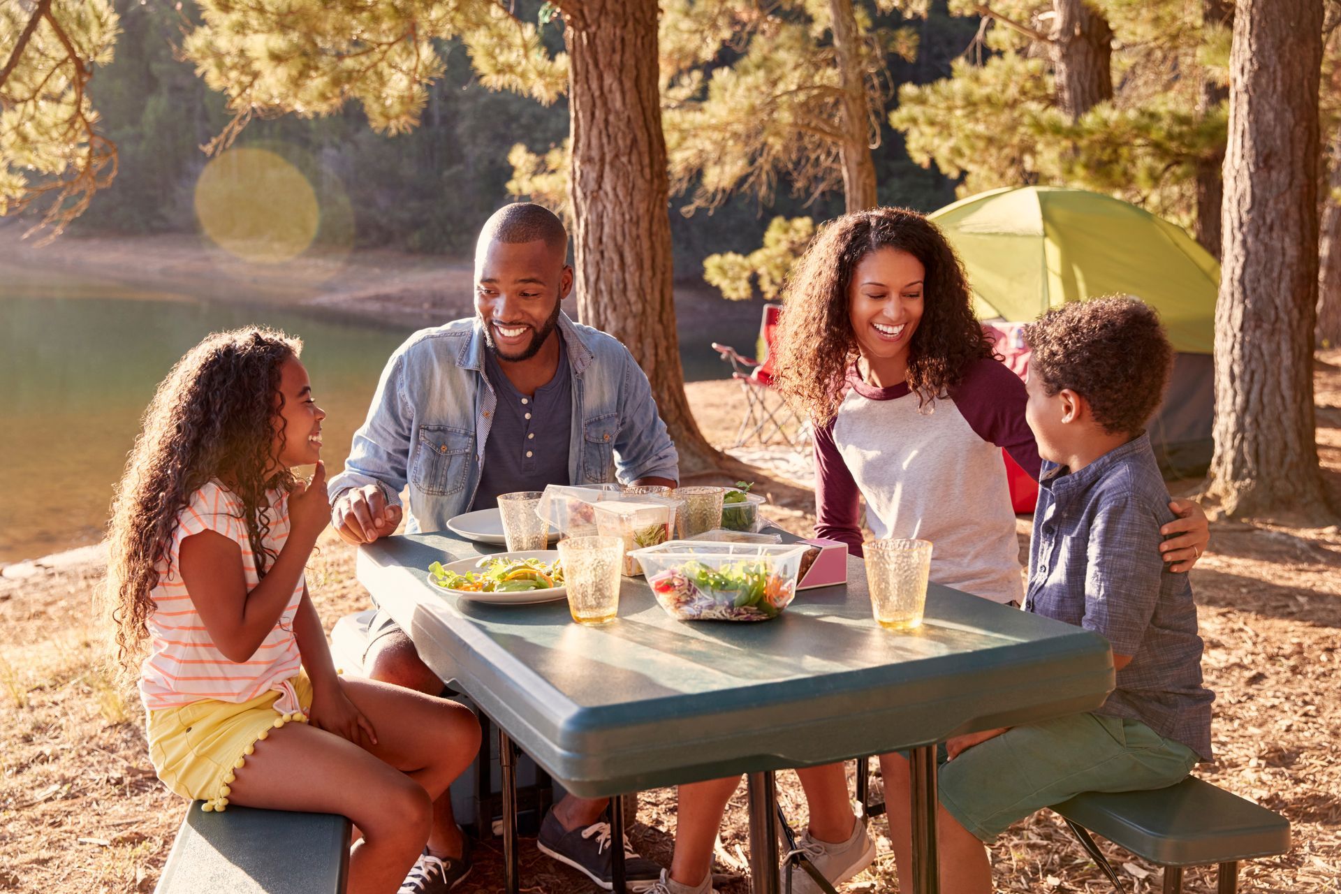 A family is sitting at a picnic table in the woods eating food.