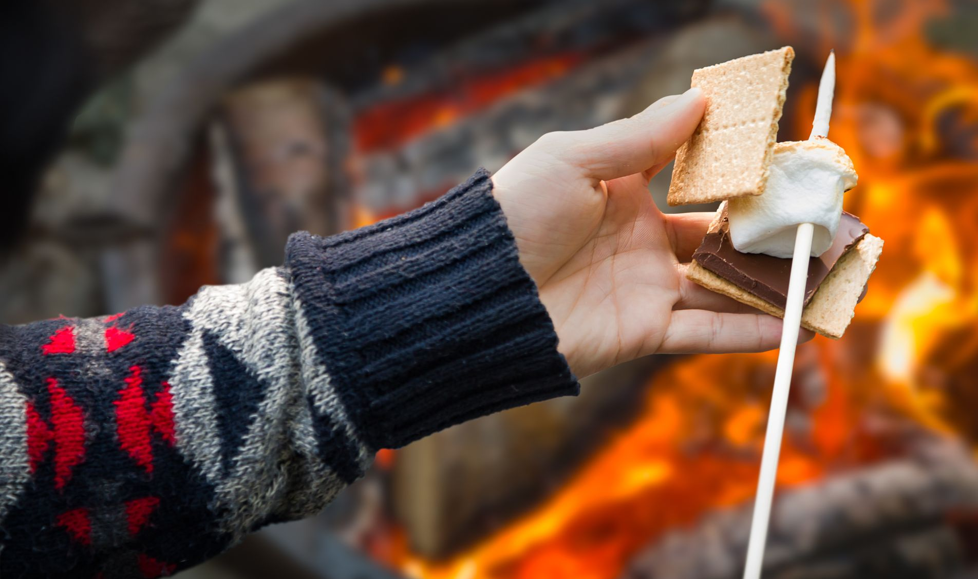 Hand holding a s'more on a stick over a campfire; sweater with red, blue, and white patterns.