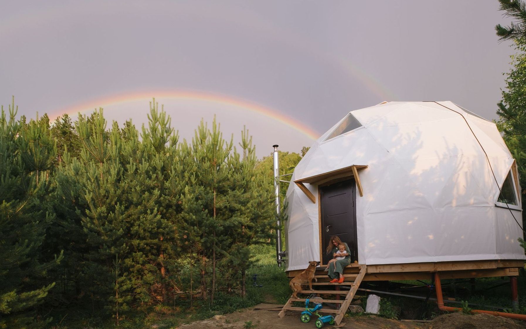 A person is sitting on the steps of a dome tent under a rainbow.