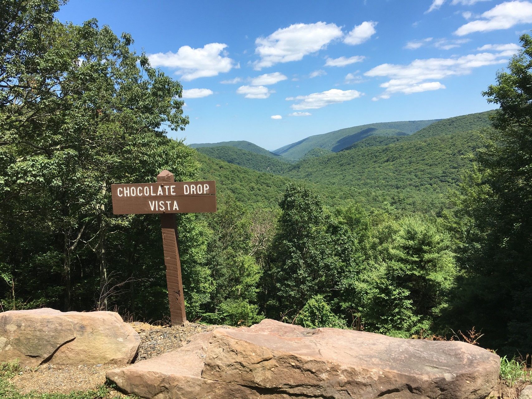 A wooden sign sitting on top of a rock overlooking a valley.