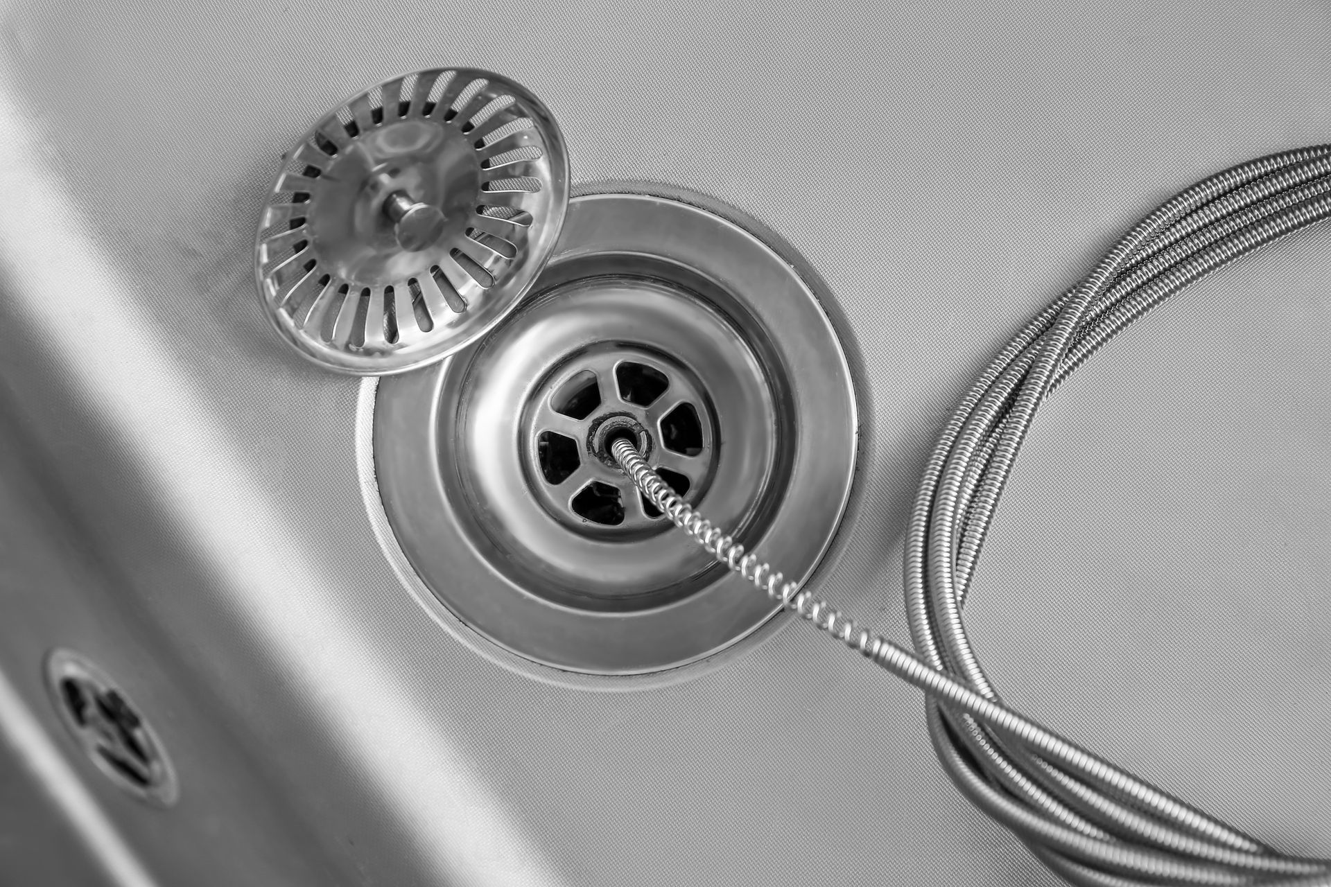 A metal drain with a hair strainer and drain snake extending into it, angled view.