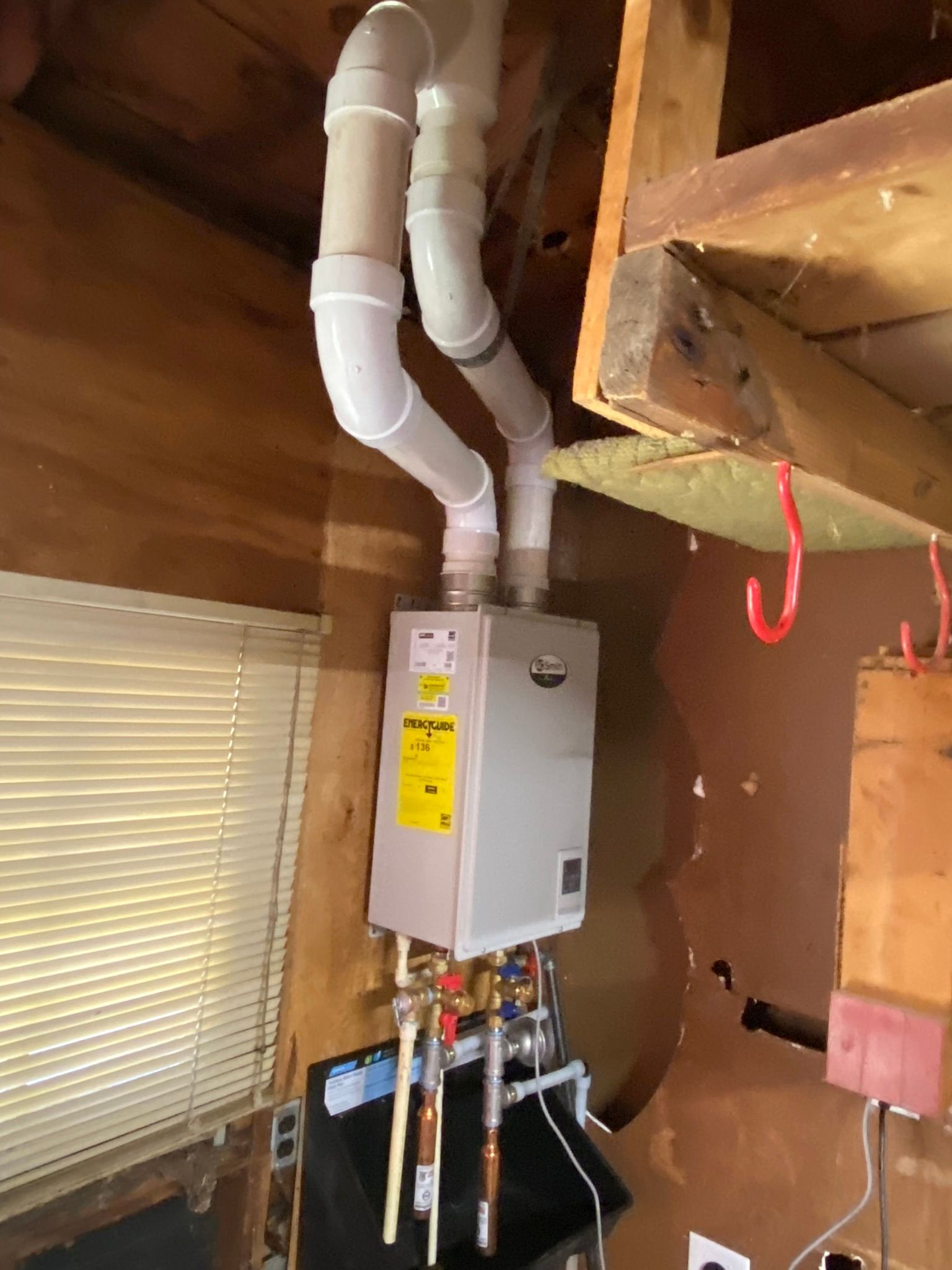 Tankless water heater installed in a wooden room, with white pipes extending upwards.