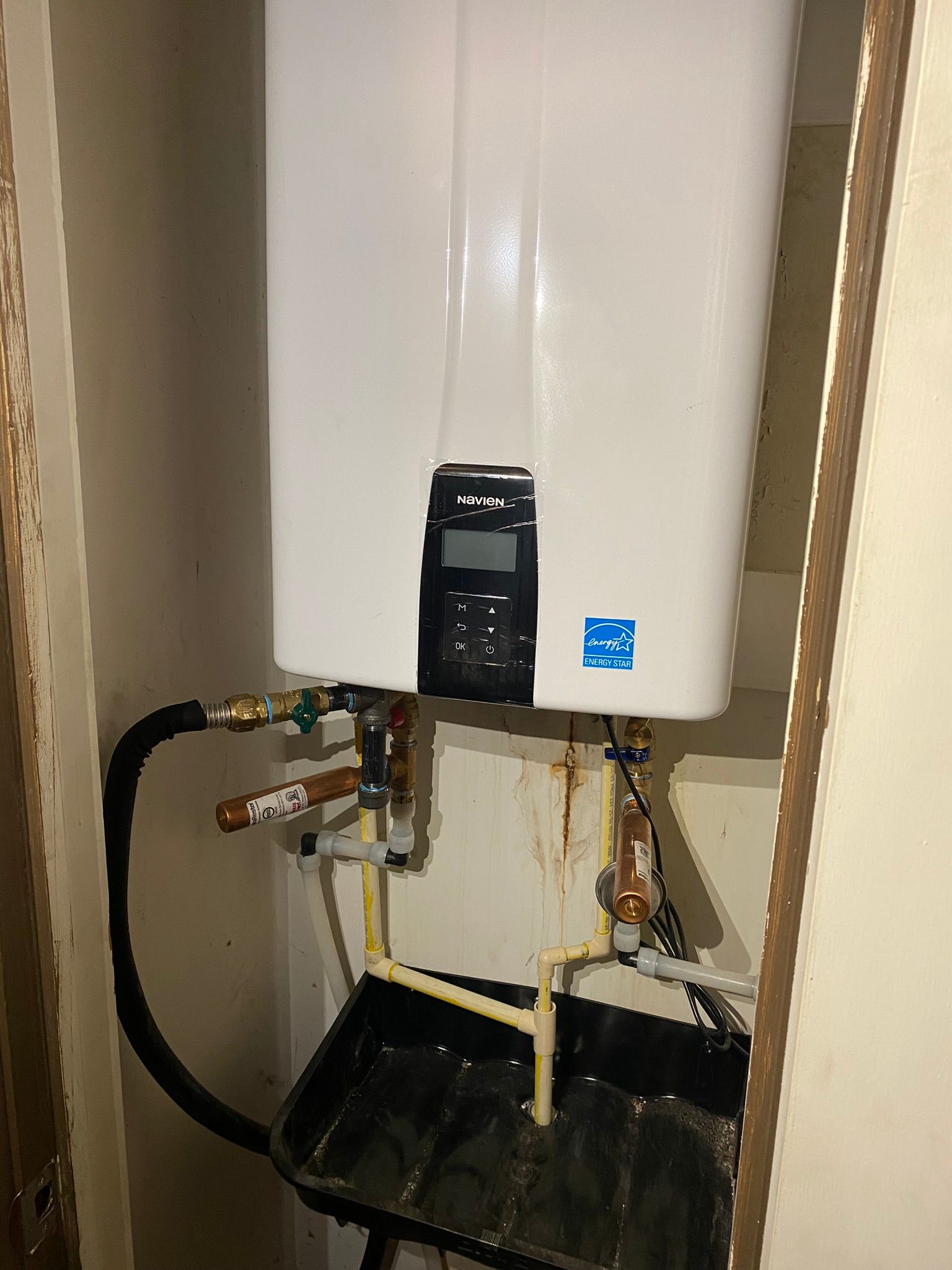 White tankless water heater mounted on a wall. Plumbing pipes and a black catch basin are below.