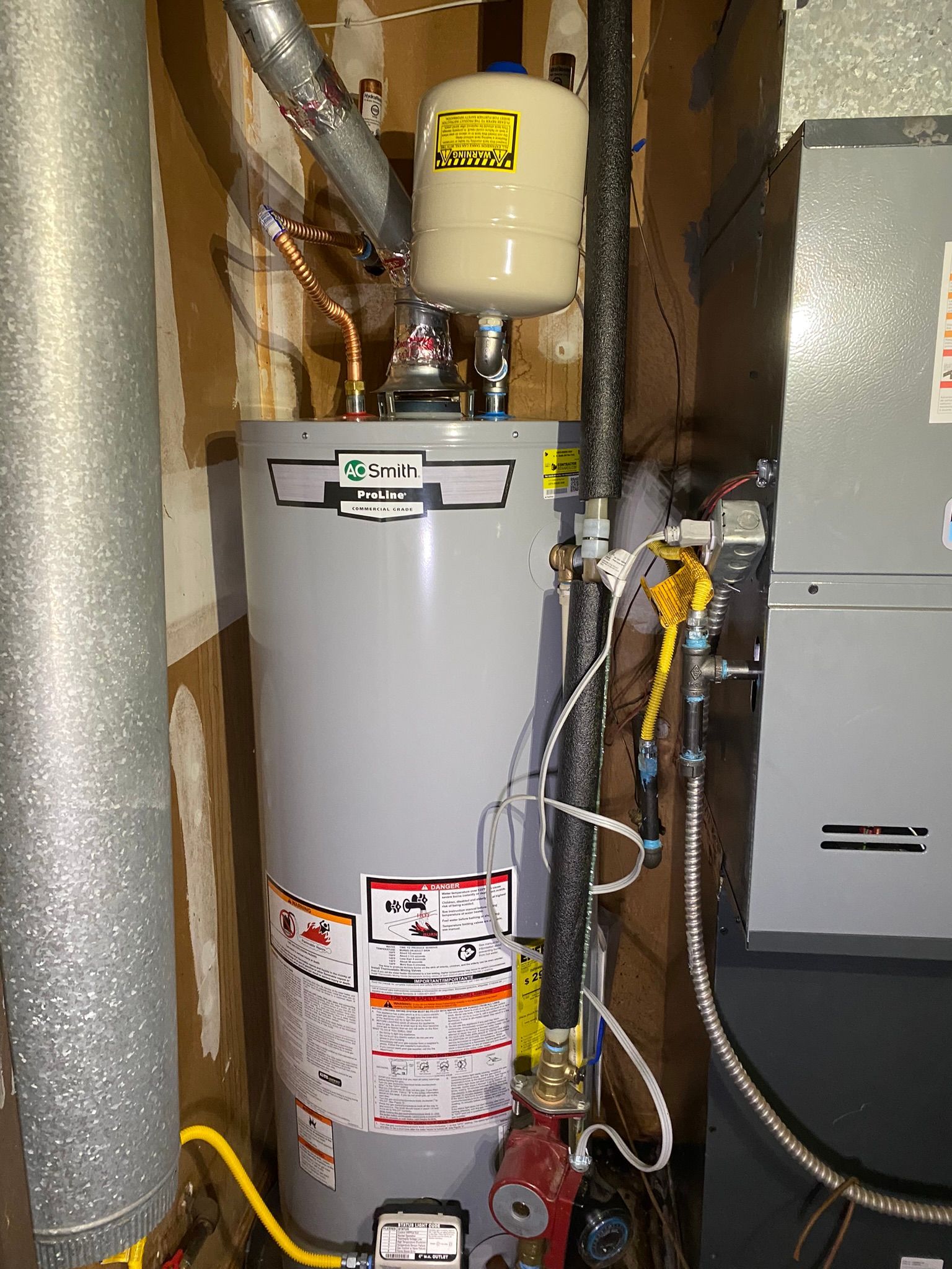 A gray water heater with a white expansion tank in a utility closet next to a furnace.