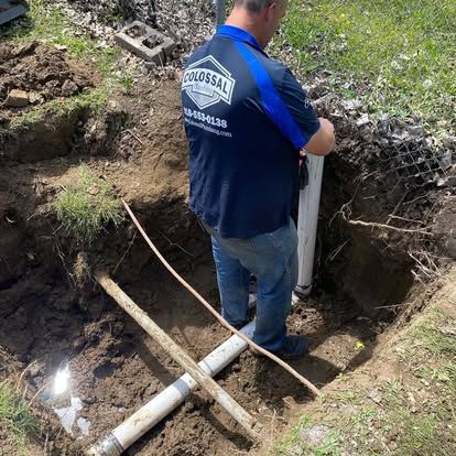Plumber in a dirt pit, working on white pipes. Blue and black logo on shirt.