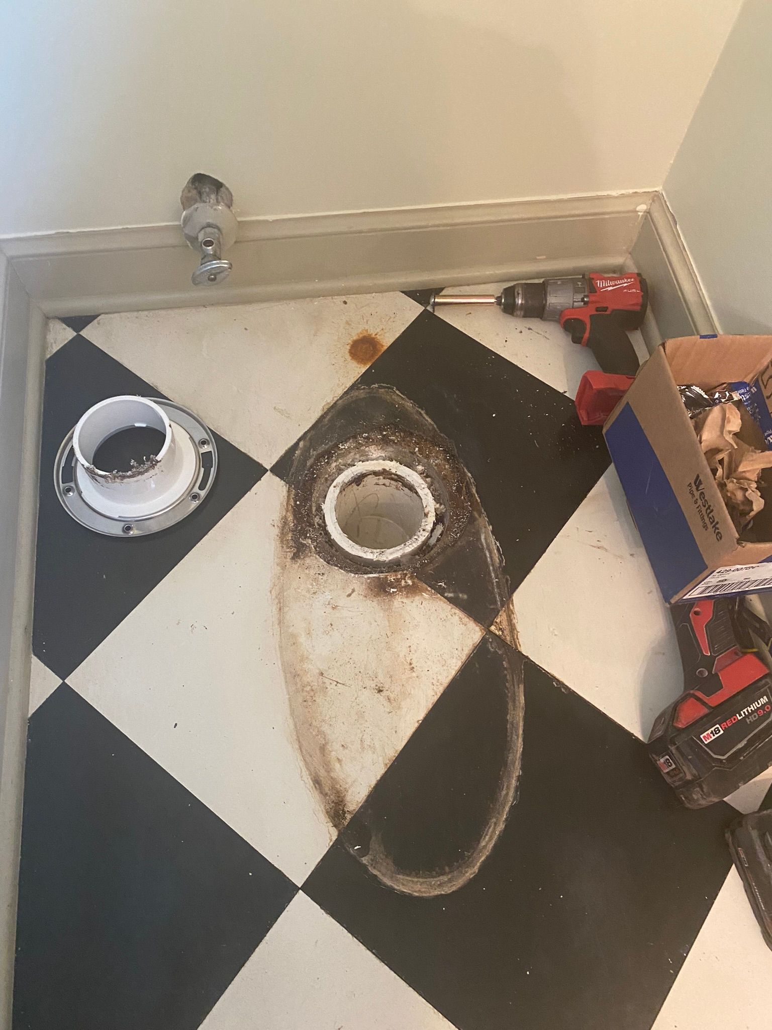 Toilet flange and drain hole visible on a checkered floor, tools nearby, likely bathroom remodel.