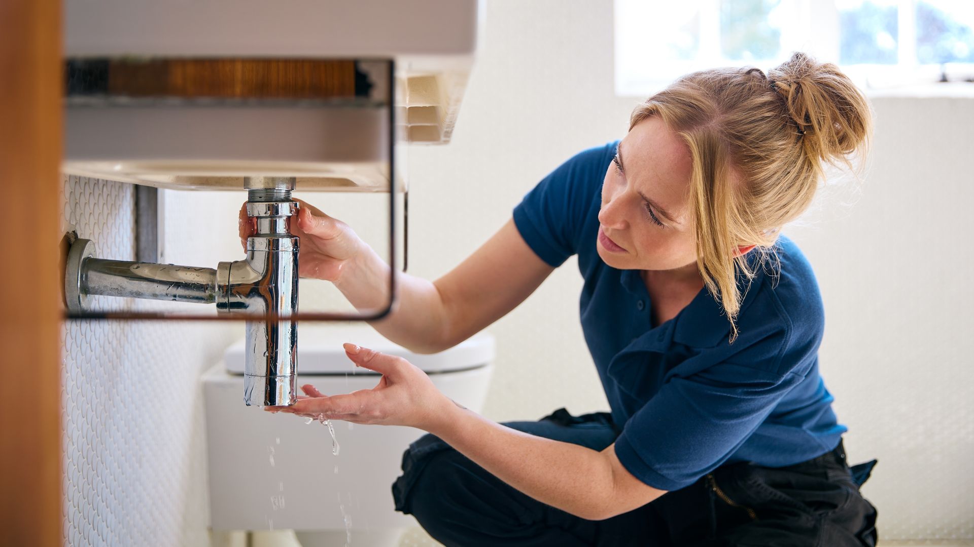 Woman in blue shirt inspecting bathroom plumbing, water dripping.