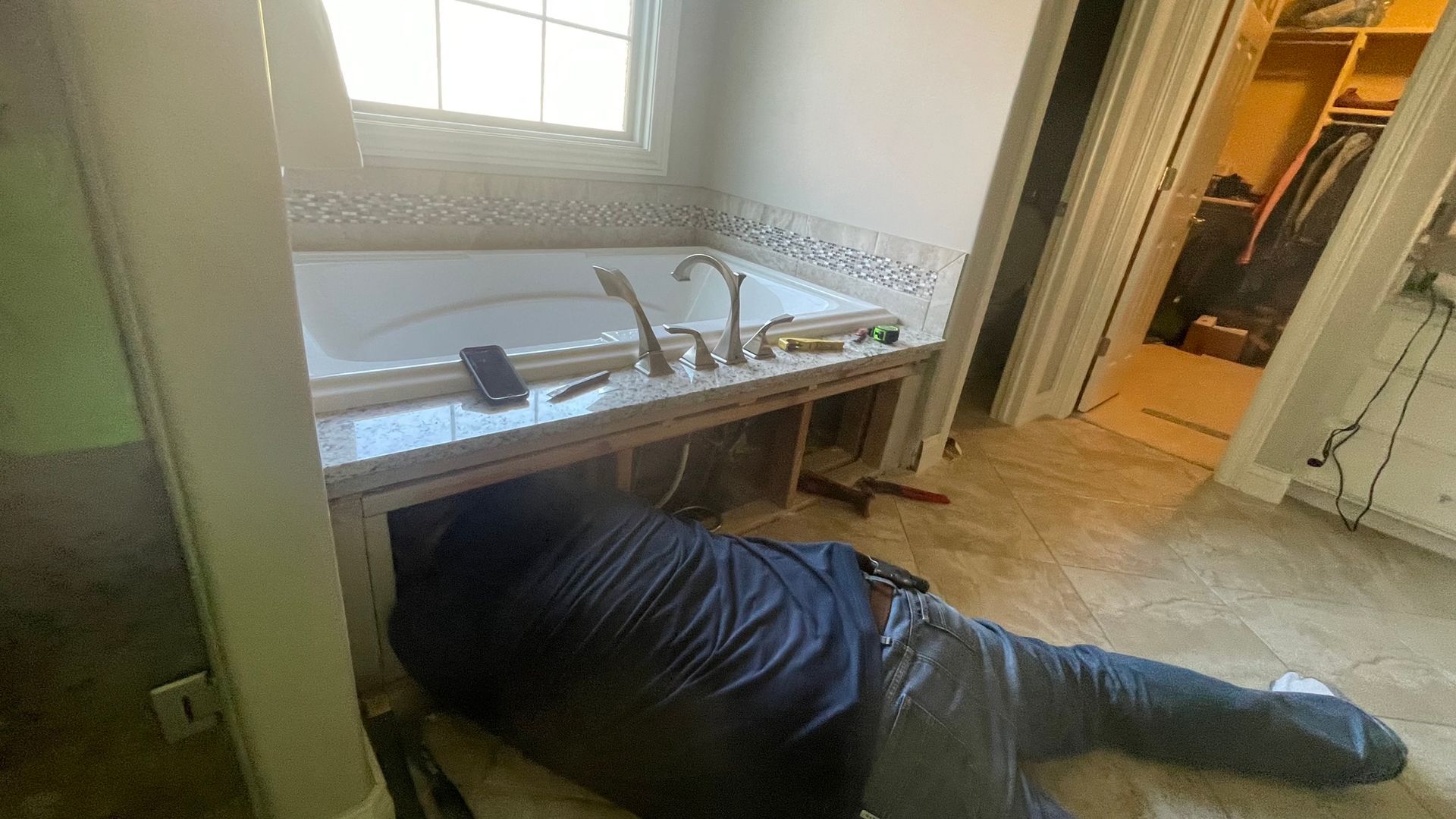 Man works under a built-in bathtub, tools and a phone nearby. Bathroom setting.